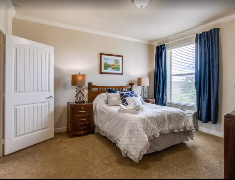 A bright, furnished bedroom with a made bed, wooden headboard, two nightstands with lamps, a window with blue curtains, and an open door.
