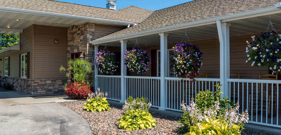 Exterior view of a senior living facility with a covered porch featuring white railings and hanging flower baskets. The building has beige siding with stone accents and a paved walkway surrounded by landscaped plants and flowers.