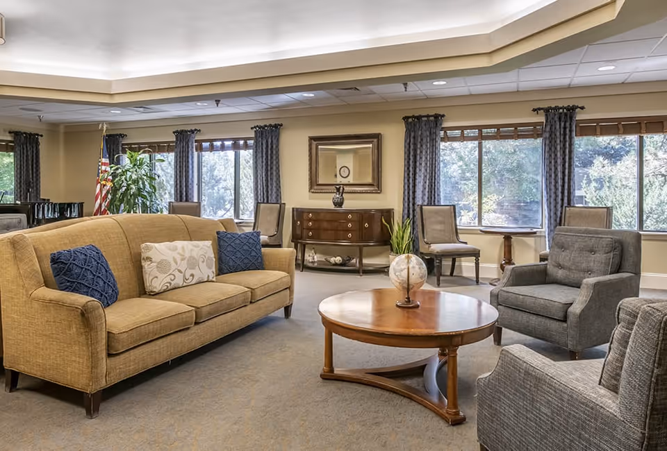 A well-lit living room with a beige sofa adorned with three decorative pillows, two blue and one patterned. There are two gray armchairs and several wooden chairs arranged around a round wooden coffee table with a small globe and plant on it. Large windows with blue curtains let in natural light, and a wooden sideboard with a mirror above it is against the far wall. The room has a neutral color palette and a cozy, inviting atmosphere.