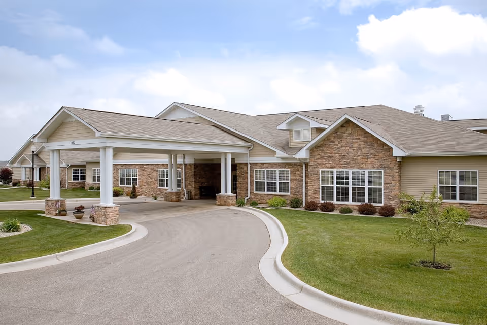 Exterior view of a single-story rehabilitation and healthcare center building with a covered entrance, stone and siding facade, multiple windows, and a curved driveway surrounded by green lawns and small plants under a partly cloudy sky.