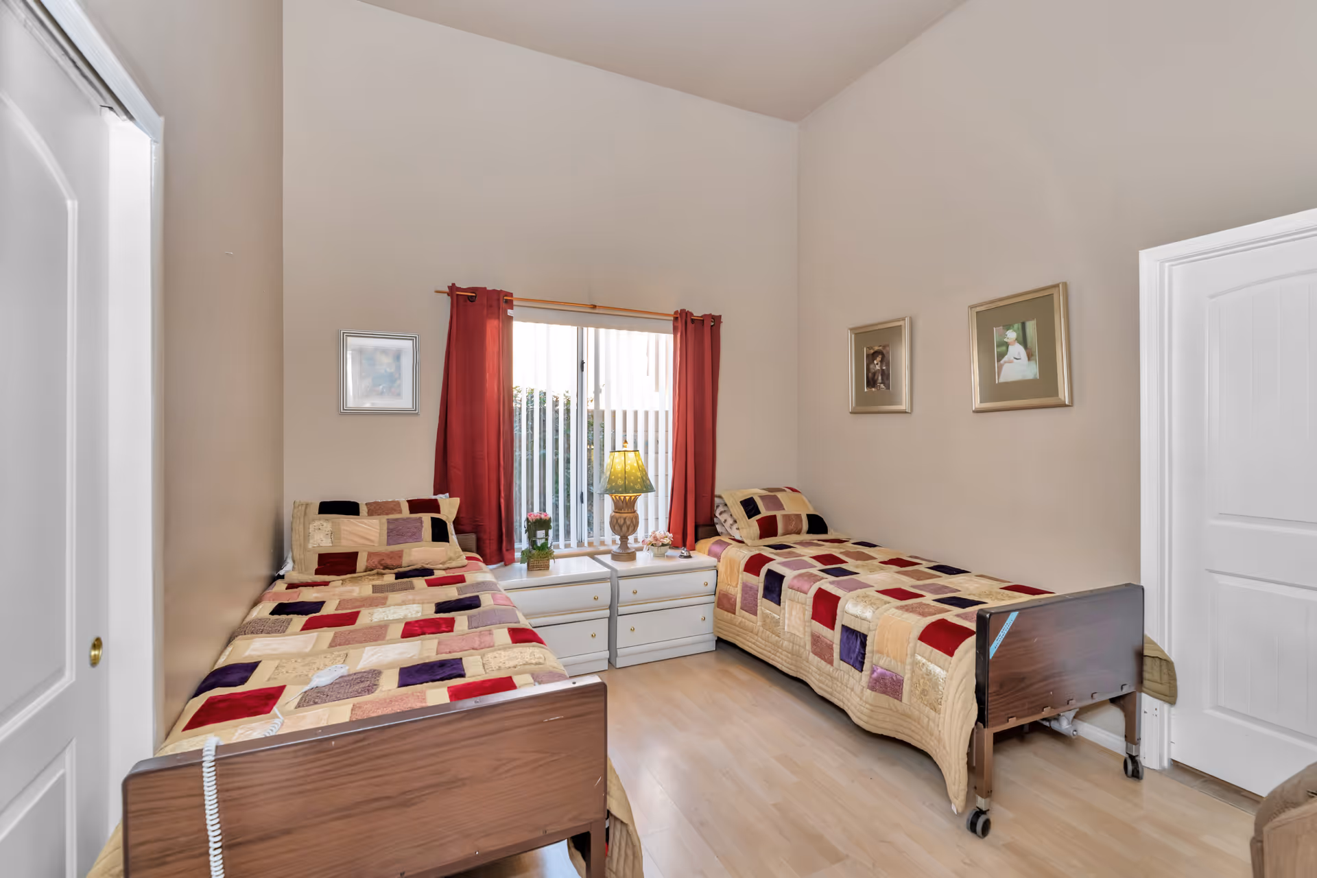 A bedroom with two single beds featuring patchwork quilts in shades of beige, red, purple, and brown. Between the beds is a white nightstand with a lamp and small decorative plants. The room has light-colored walls, a window with vertical blinds and red curtains, and framed pictures on the walls. The floor is light wood, and one bed has a call button cord attached.