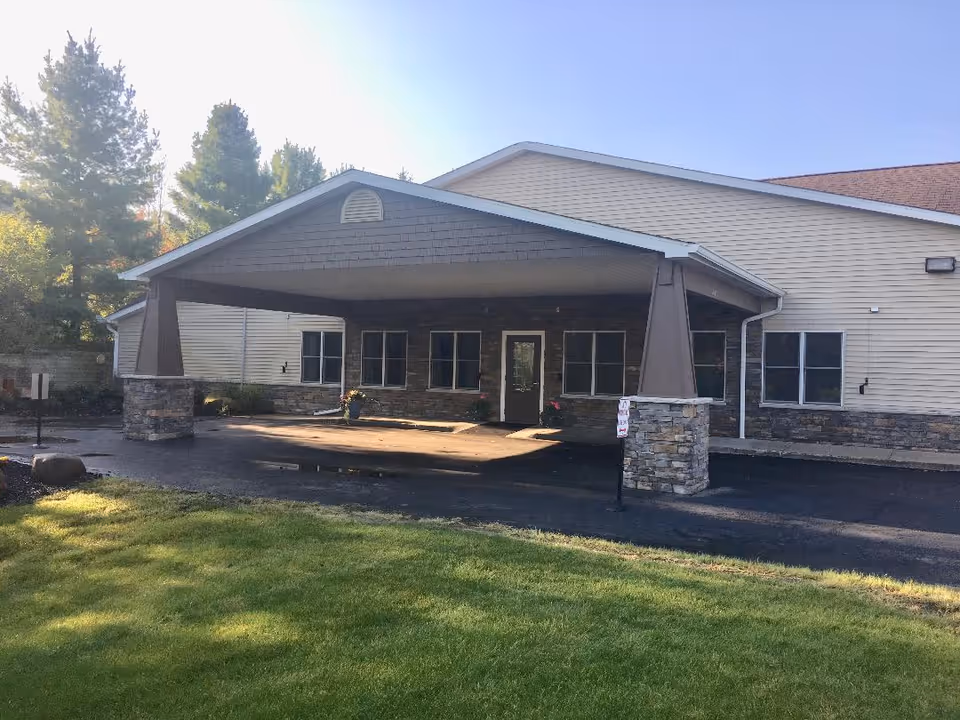 Exterior view of a single-story building with a covered entrance supported by stone pillars. The building has beige siding and multiple windows, with a paved driveway and green grass in the foreground. Trees are visible in the background under a clear sky.