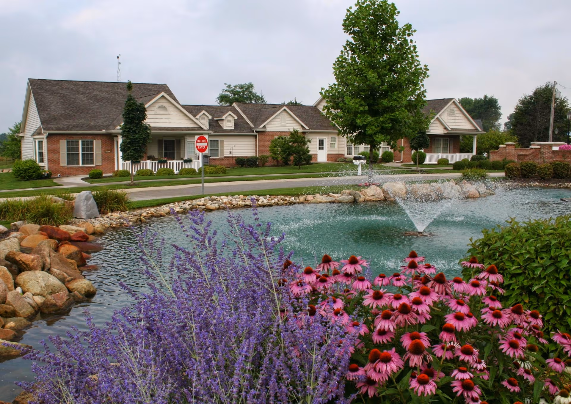 A landscaped pond with a fountain and blooming purple and pink flowers in front of single-story brick-and-siding residential buildings.