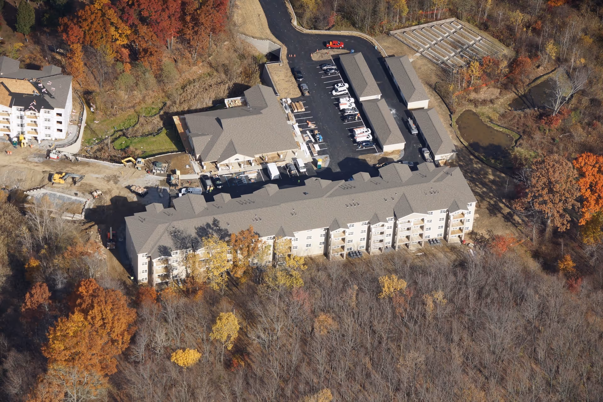 Aerial view of Glenmont Abbey Village senior living facility surrounded by autumn trees with colorful foliage. The image shows multiple buildings, parking areas with vehicles, and some construction equipment. There is a small pond visible on the right side and a wooded area in the foreground.
