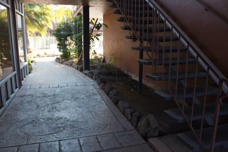 A shaded outdoor walkway with textured concrete paving leading past a garden bed with small plants and rocks on the right, and a metal staircase with open steps ascending along a brown wall.