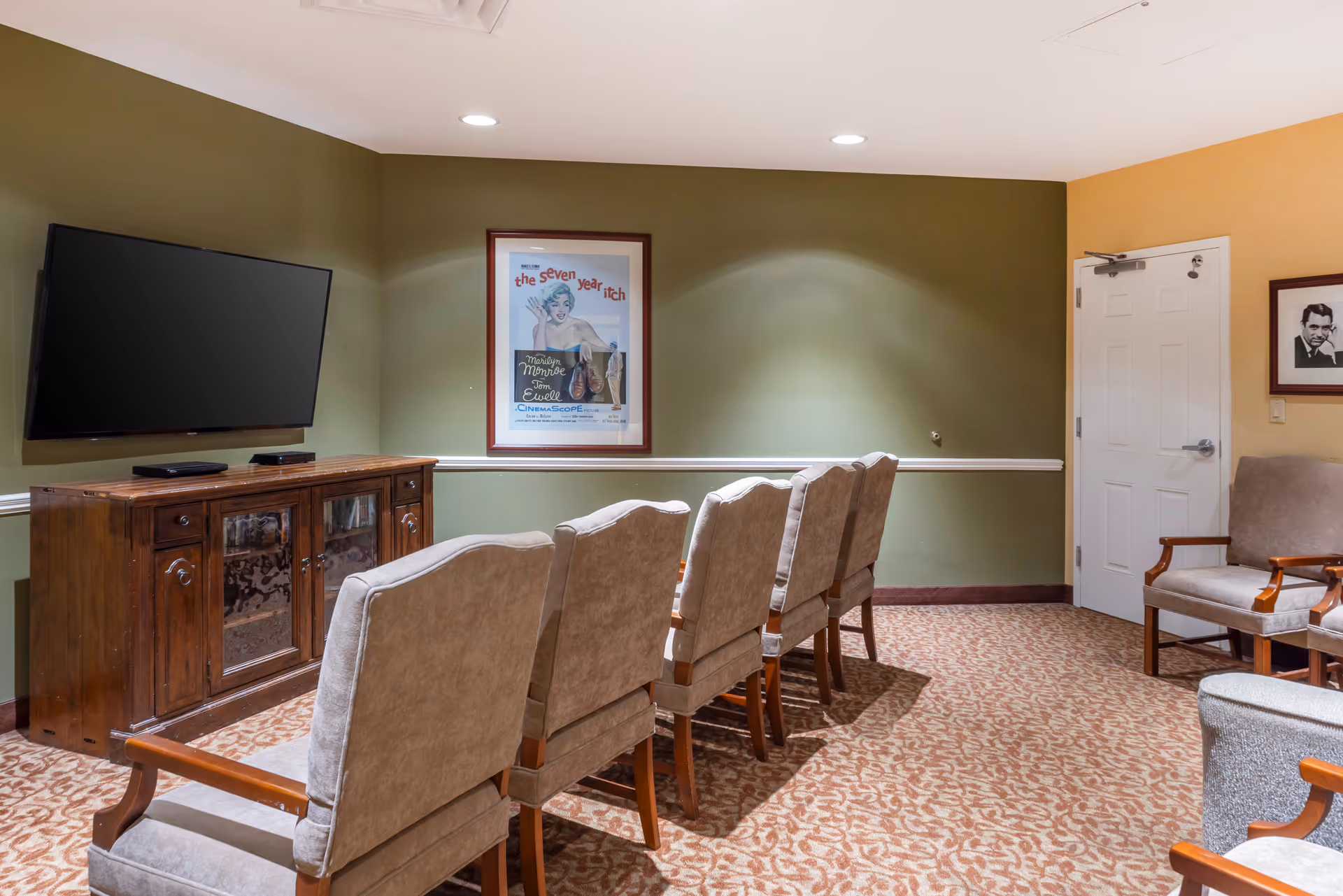 A small media room with six beige upholstered chairs arranged in two rows facing a wall-mounted flat screen TV above a wooden cabinet. The walls are painted green and yellow, with a framed vintage movie poster and a black and white portrait hanging on the walls. The floor is carpeted with a patterned design.
