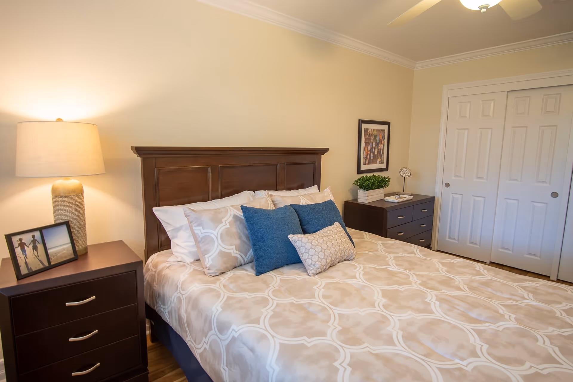 A neatly made bed with beige and white patterned bedding and blue accent pillows in a bedroom. On the left side, there is a dark wooden nightstand with a lamp and a framed photo. On the right side, there is a matching dresser with a small plant and decorative items. The room has beige walls, white closet doors, and a ceiling fan.