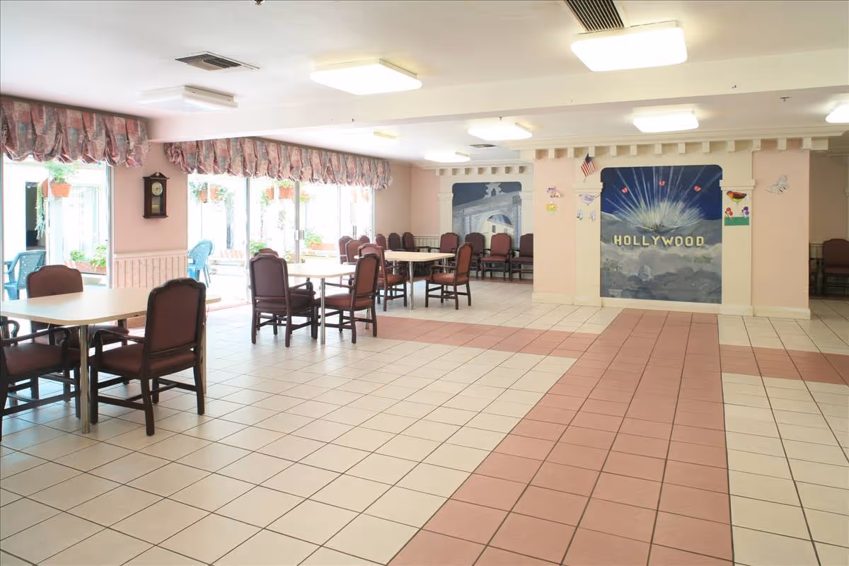 A spacious dining area with several tables and chairs arranged neatly. Large windows on the left side let in natural light and have floral valances. The far wall features murals, including one with the word 'HOLLYWOOD' and decorative butterflies and flowers. The floor is tiled with a pattern of white and pinkish tiles.