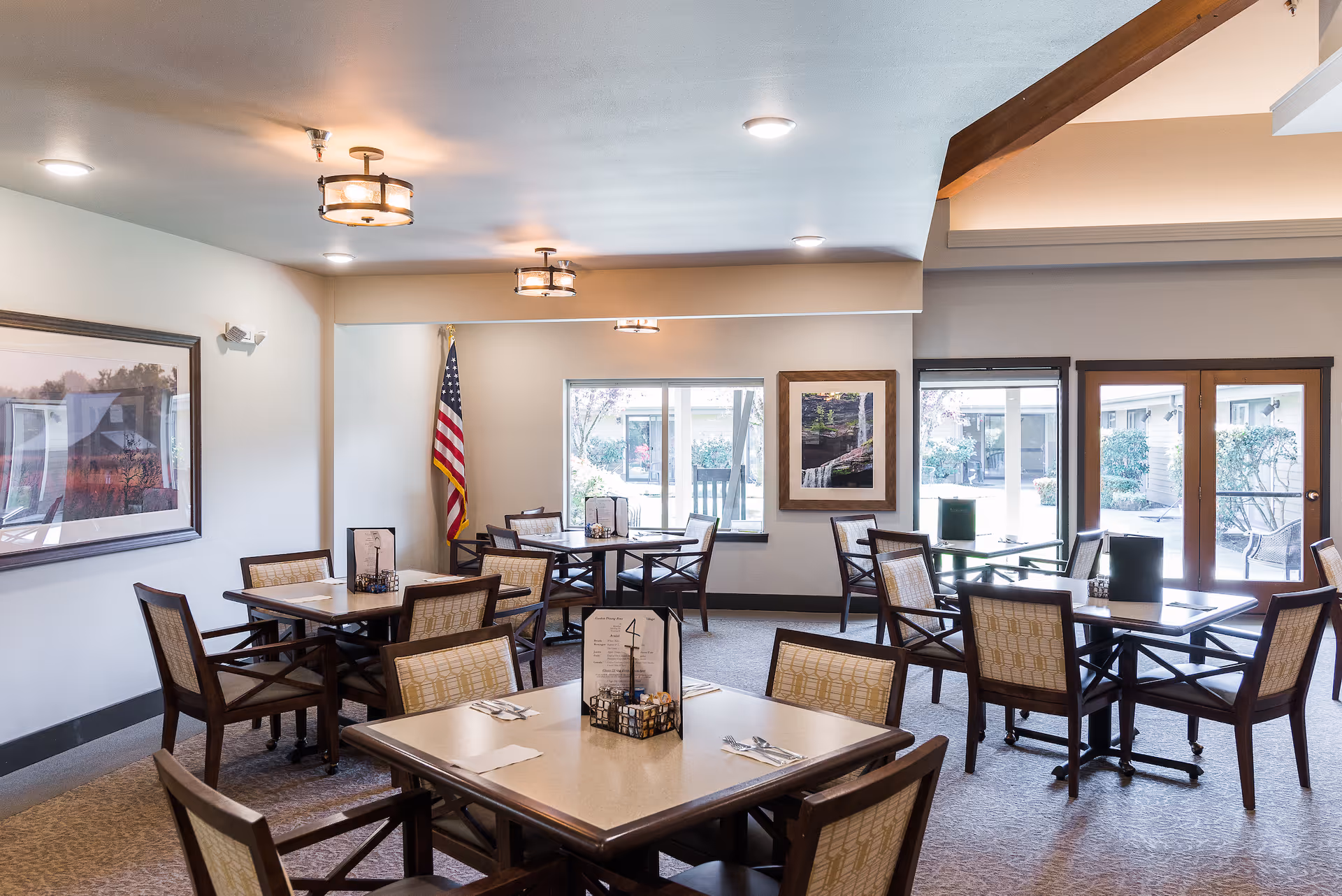 A dining room in a senior living facility with several square tables and chairs arranged neatly. The room has large windows and glass doors letting in natural light, framed artwork on the walls, and an American flag in the corner. The ceiling has modern light fixtures and the floor is carpeted.