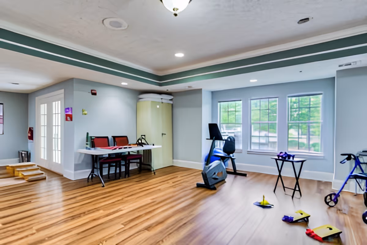 A spacious room with wooden flooring and light blue walls featuring a stationary exercise bike, a walker, a small table with purple dumbbells, and a folding table with chairs. Large windows allow natural light to enter, and there is a set of French doors on the left side.