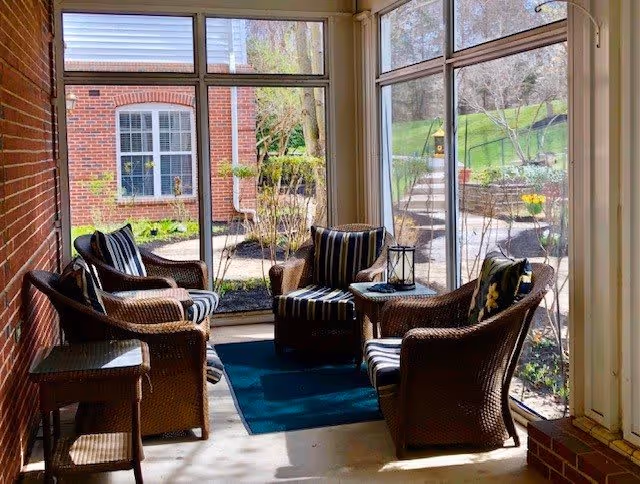 A sunlit indoor seating area with four wicker chairs arranged around a small table, all with striped cushions. The space has large windows providing a view of a garden and brick building outside. A blue rug is placed under the table and chairs.