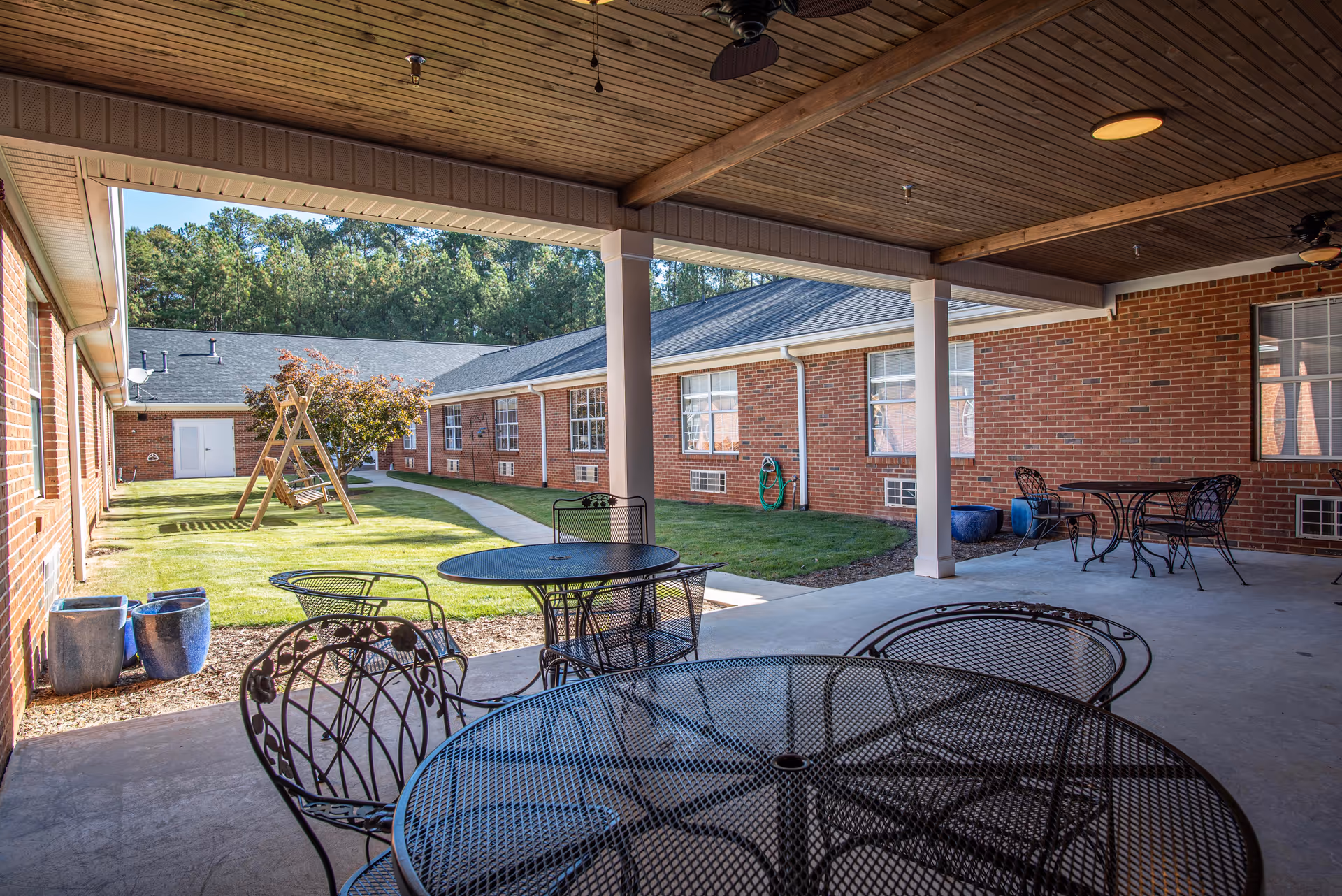Covered outdoor patio area with metal tables and chairs overlooking a grassy courtyard with a wooden swing set and brick building walls surrounding the space.