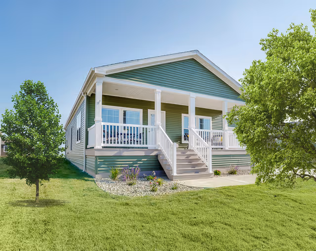 Exterior view of a single-story green house with white trim and a front porch with railings and stairs leading up to the entrance. The house is surrounded by a well-maintained lawn with two trees on either side and a clear blue sky overhead.