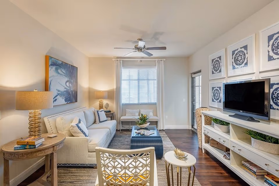 Well-lit living room with a white sofa, coffee table, TV on a console, and framed wall art.