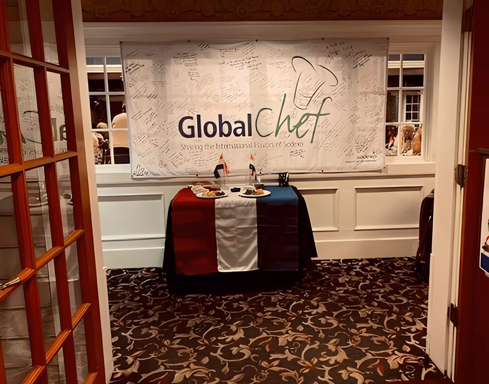 A small table covered with a cloth in the colors of the French flag (blue, white, and red) is set up in a room with patterned carpet and white paneled walls. On the table are two plates of food and two small flags. Behind the table is a large banner that reads 'Global Chef' with a chef's hat graphic and additional text below. The room is viewed through an open door with glass panes.