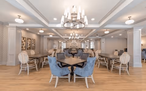 A spacious dining room with multiple tables and chairs arranged neatly. The room features elegant chandeliers and ceiling lights, light-colored walls with decorative molding, and framed artwork on the walls. The floor is wooden, and the overall ambiance is bright and welcoming.