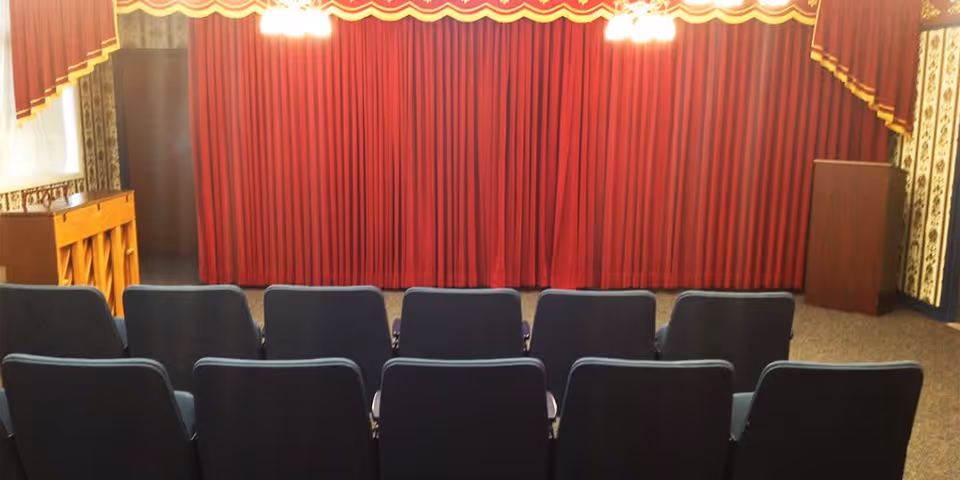 Small auditorium or theater room with two rows of blue chairs facing a stage with closed red curtains. There is a wooden piano on the left side and a wooden podium on the right side. The room has patterned wallpaper and ceiling lights above the stage.