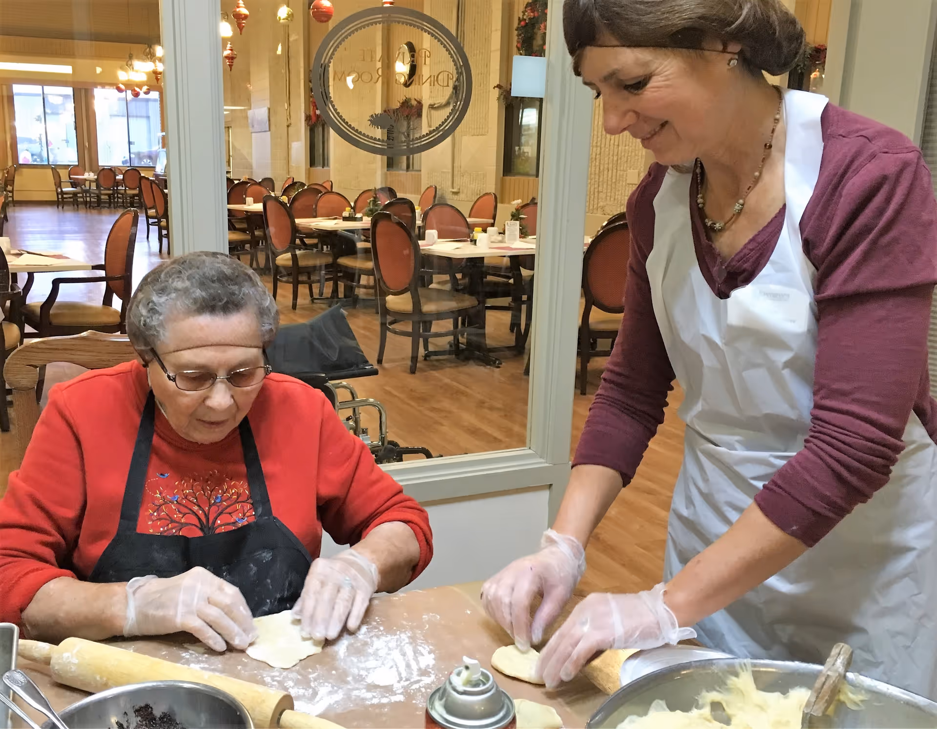 Two women wearing aprons and gloves roll out dough on a table in a communal dining area with empty tables visible through a glass partition.