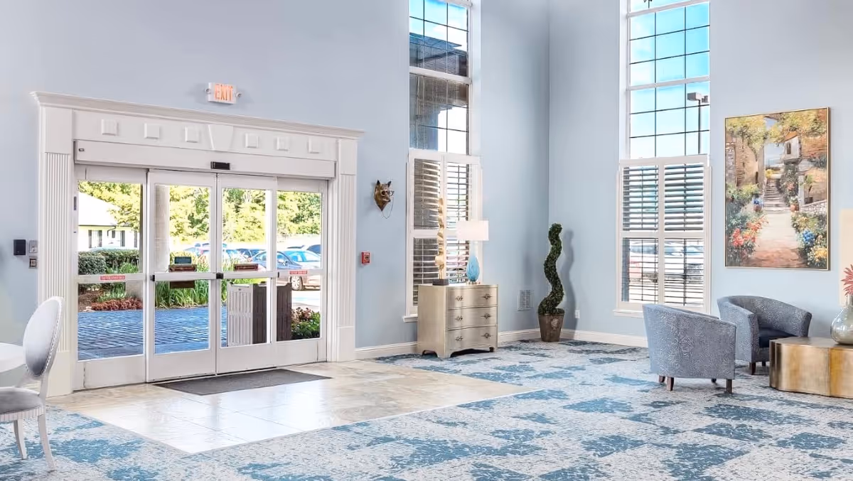 Bright reception lobby with glass double doors, tall windows, patterned carpet, and a seating area with chairs and decorative furnishings.