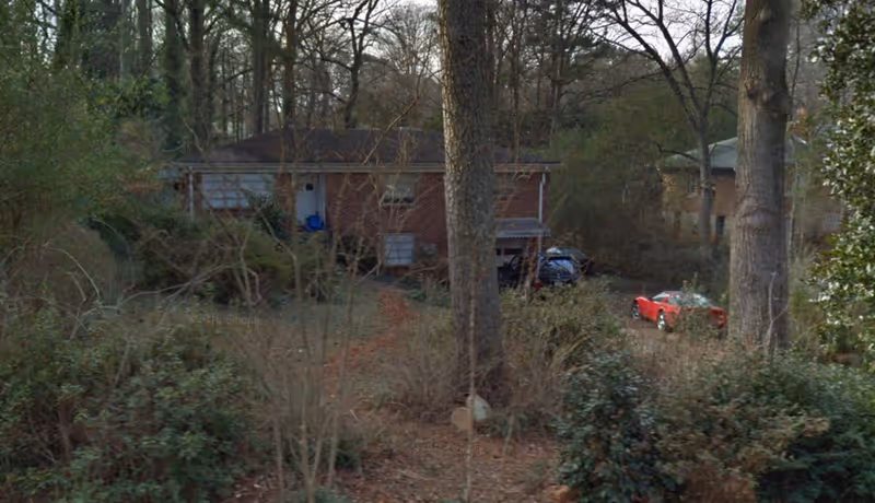 Front view of a brick house partially obscured by trees and overgrown yard with two parked cars.