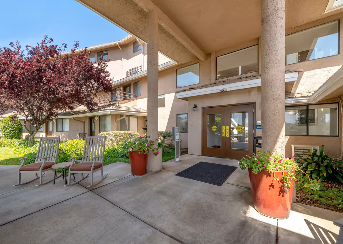 Entrance area of Las Brisas Retirement Community showing a covered walkway with two large columns, two wooden rocking chairs with cushions, a small table, large red flower pots with green plants and flowers, and a beige multi-story building with balconies and windows in the background under a clear blue sky.