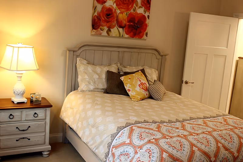 Neatly made bed with patterned bedding and decorative pillows, a nightstand with a lamp, and floral artwork on the wall in a cozy bedroom.
