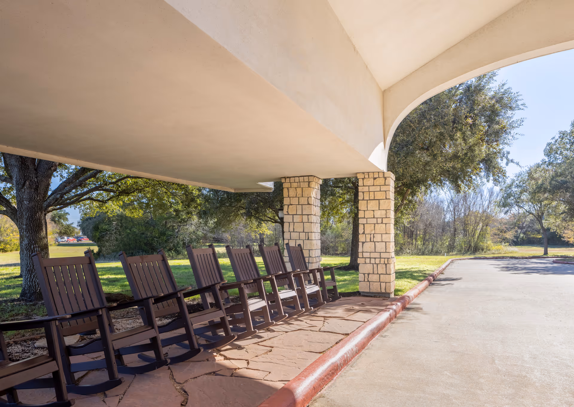 Covered front entrance with a row of wooden rocking chairs under a wide overhang supported by stone pillars, facing a driveway and trees.