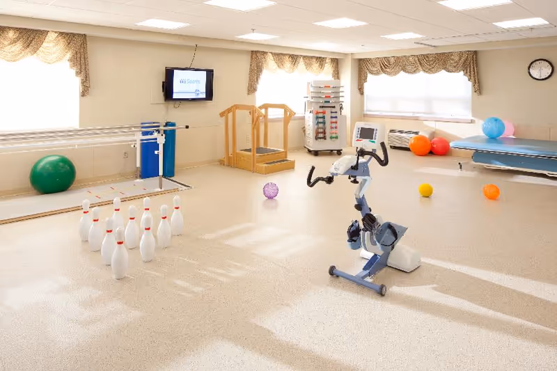 Bright rehabilitation/exercise room with a recumbent exercise bike, parallel bars, therapy table, colorful exercise balls and a set of plastic bowling pins.