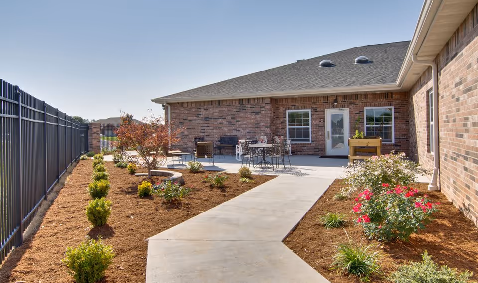 Outdoor patio area at Lakewood Senior Living featuring a concrete walkway leading to a seating area with metal tables and chairs. The patio is surrounded by landscaped garden beds with small shrubs, flowering plants, and a tree. The building has brick exterior walls and several windows, with a clear blue sky overhead.