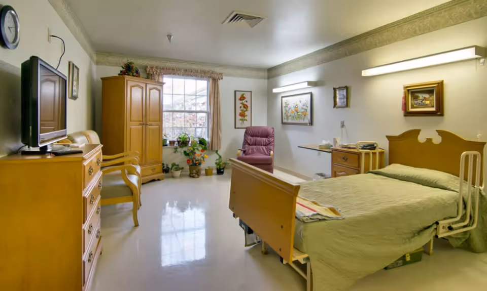A well-lit skilled nursing facility room with a hospital-style bed covered in a green quilt, a wooden dresser with a TV on top, two wooden chairs, a wooden wardrobe, a purple recliner chair, several framed floral artworks on the walls, and a window with curtains and potted plants on the windowsill.