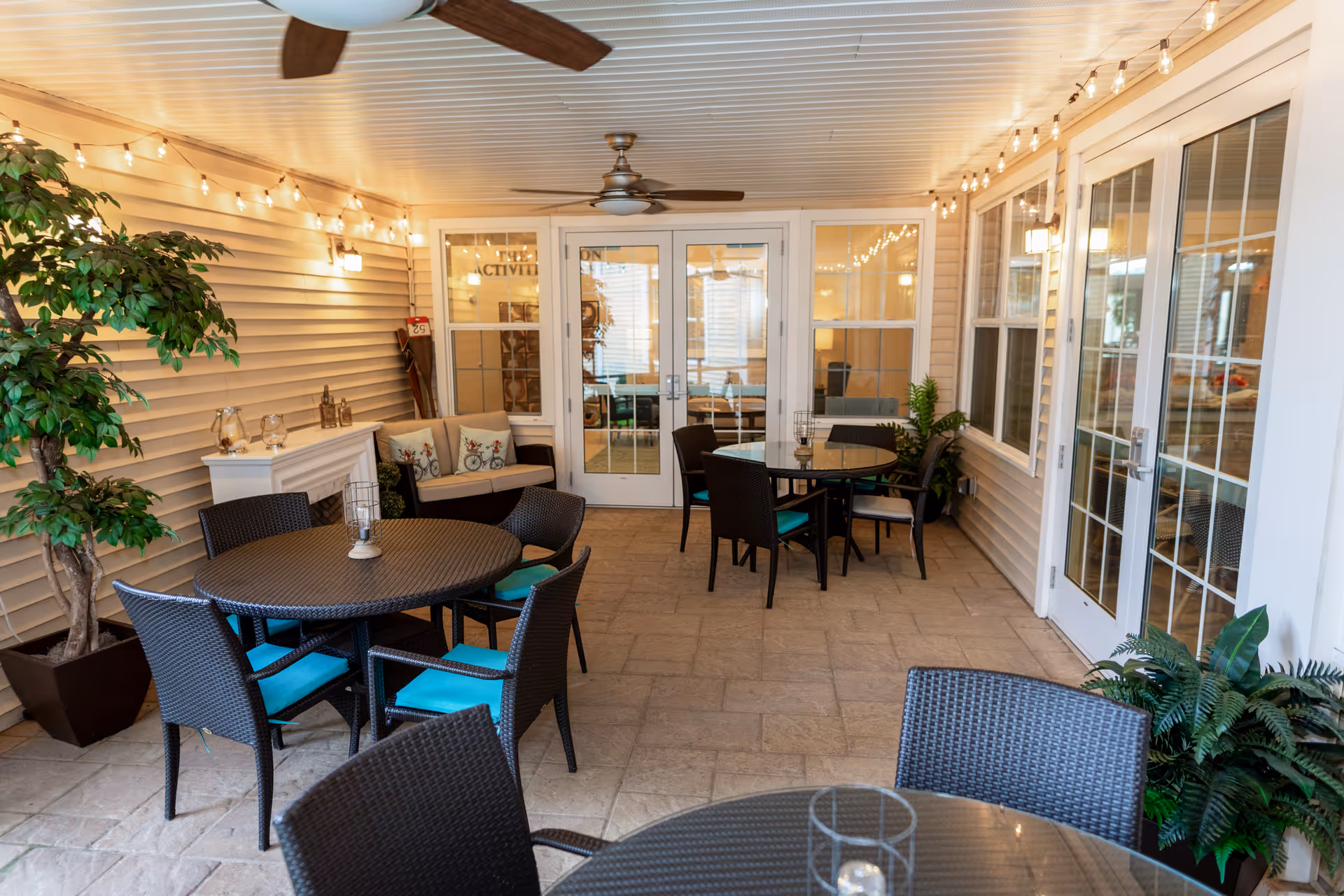 A cozy indoor patio area with round tables and wicker chairs featuring turquoise cushions. The space is decorated with string lights along the ceiling edges, potted plants, and a small white fireplace with decorative items. There are ceiling fans, large windows, and glass doors leading to another room labeled 'ACTIVITIES'.