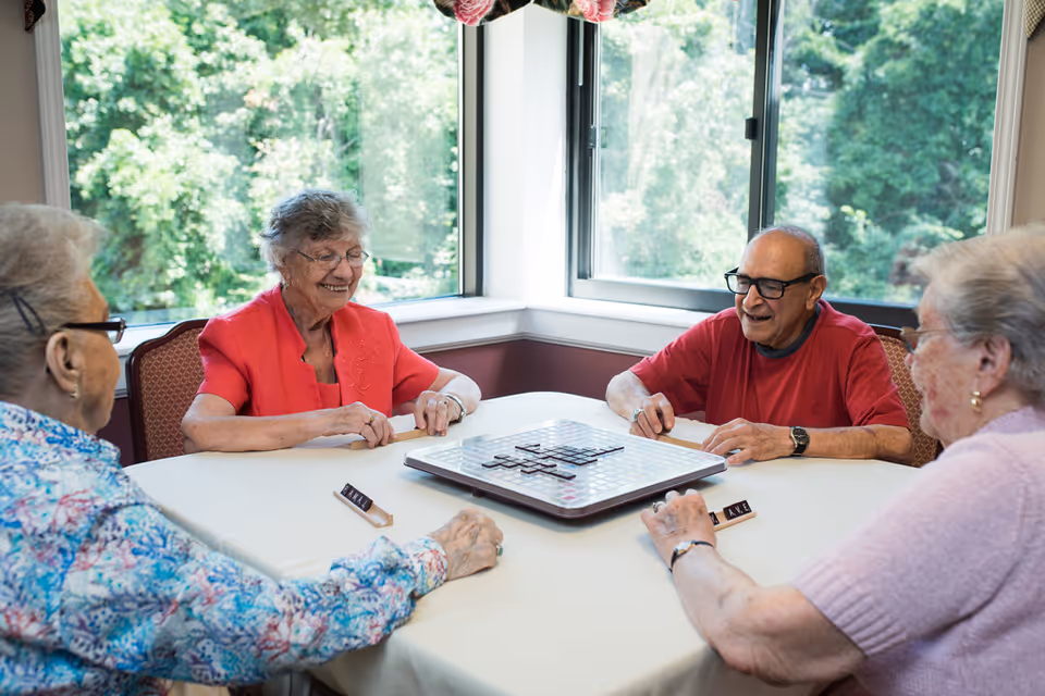 Four elderly people sitting around a table playing a board game in a bright room with large windows showing green trees outside. They appear to be enjoying the game and each other's company.