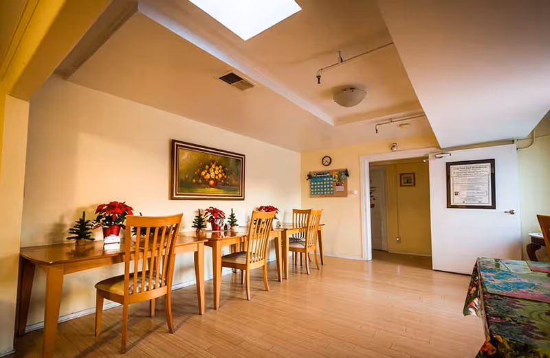 A dining area with wooden tables and chairs arranged along a wall. The tables have small decorative plants and poinsettia flowers. A framed painting of flowers hangs on the wall above the tables. The room has light-colored walls, a wooden floor, a skylight, and a door with a posted notice.