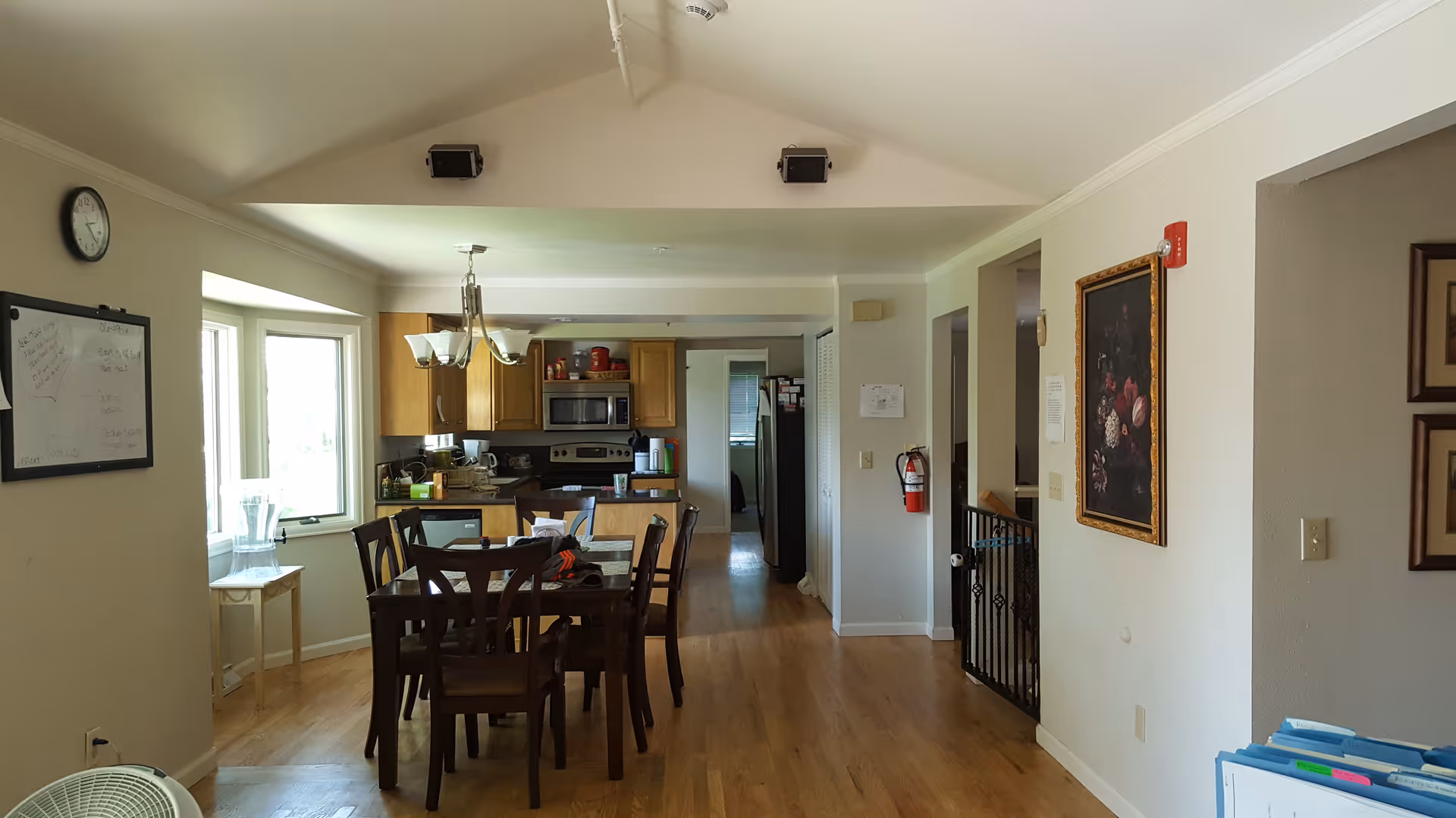 Interior view of a dining area and kitchen in an assisted living facility. The dining area features a wooden table with six chairs under a chandelier. The kitchen has wooden cabinets, a microwave, stove, and refrigerator. The room has hardwood floors, a whiteboard on the wall, a clock, and framed artwork. There is a fire extinguisher mounted on the wall near a hallway.