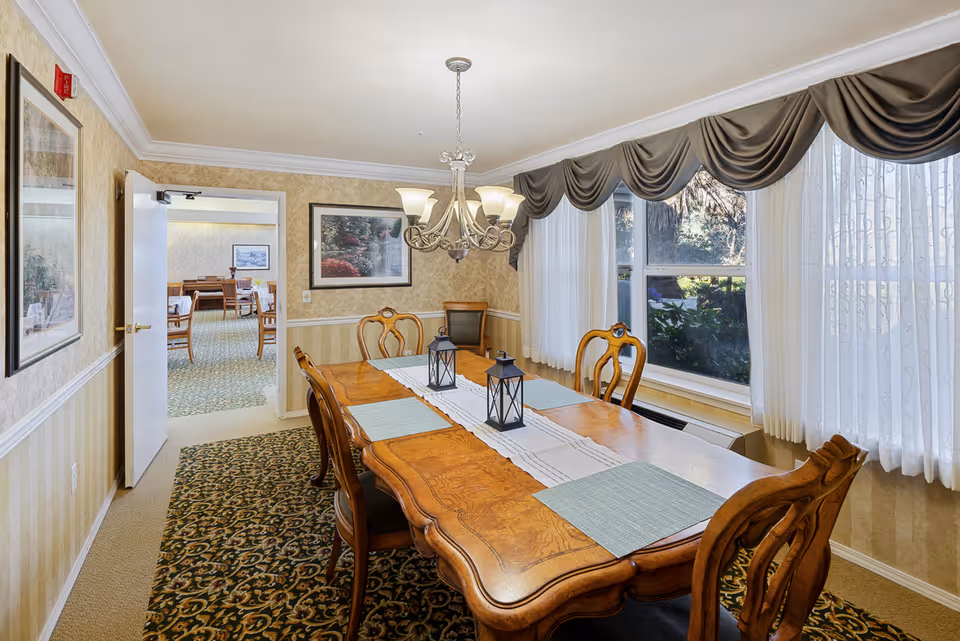 Formal dining room with a wooden table, ornate chairs, a chandelier, and large window with draped curtains.