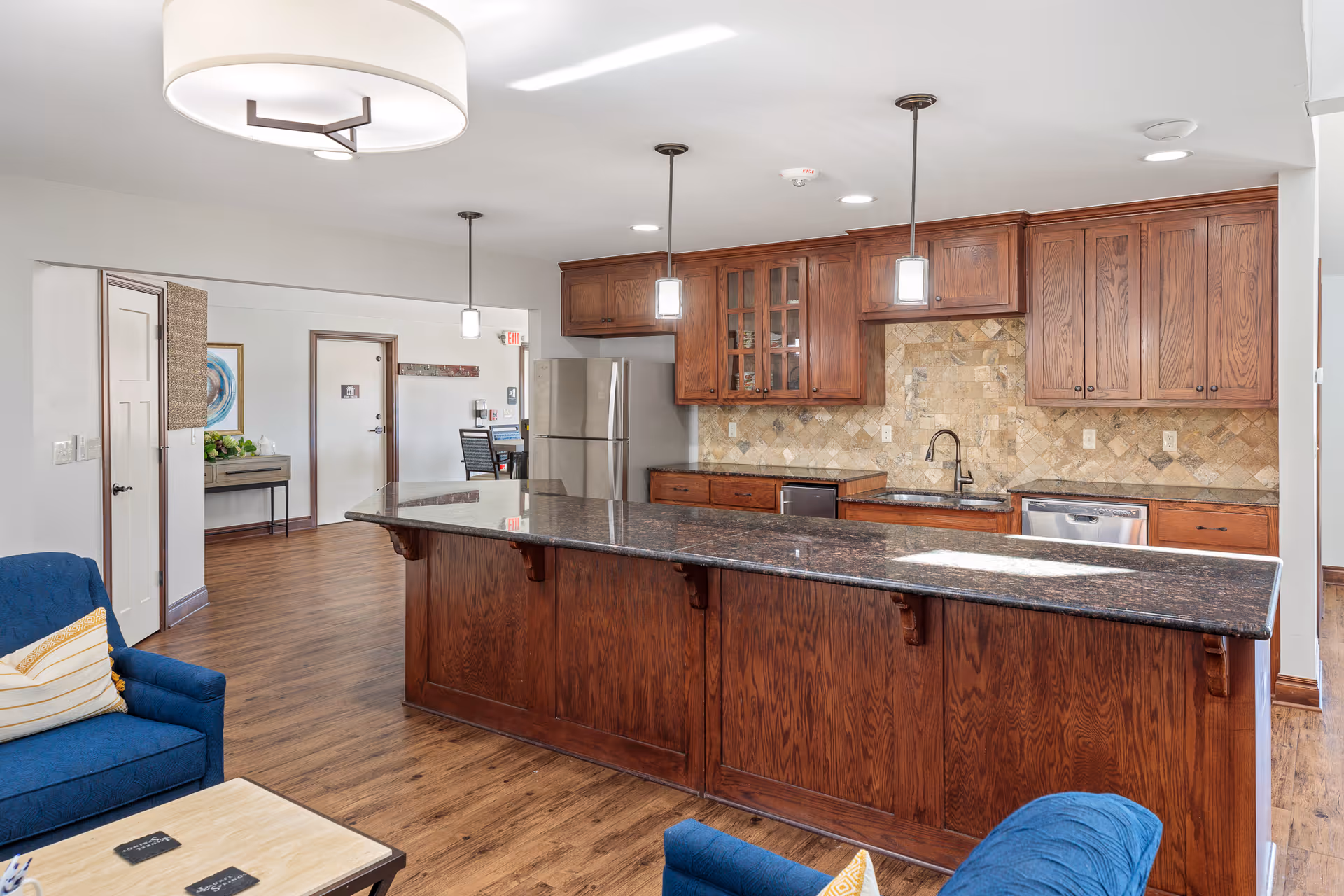 Interior view of a kitchen area in a senior living facility named Laurel Springs, featuring wooden cabinets, a granite countertop island with three pendant lights above, stainless steel refrigerator and dishwasher, and a blue armchair with a pillow in the foreground. The floor is wooden, and there is a small table with coasters near the armchair. In the background, there is a hallway with a door and some wall decorations.