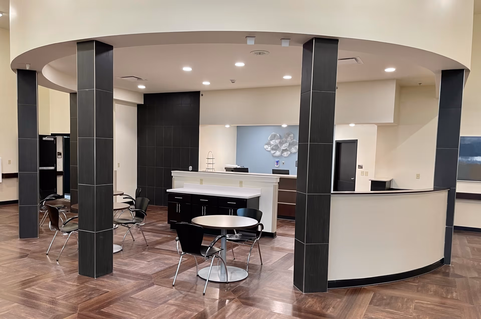 Interior view of a modern common area with a curved reception desk, dark tiled columns, round tables with black chairs, and wood-patterned flooring under bright ceiling lights.