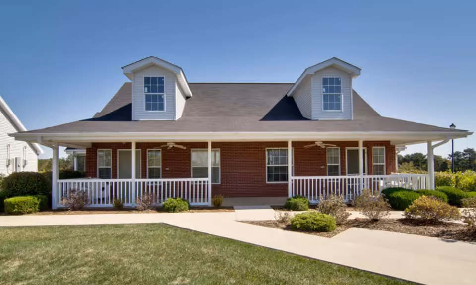 Front exterior view of a single-story brick building with two dormer windows on the roof, a covered porch with white railings and ceiling fans, surrounded by landscaped bushes and a concrete walkway under a clear blue sky.