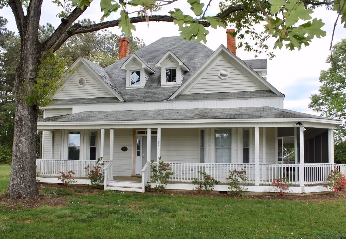 A white two-story house with a wrap-around porch, several windows, and two chimneys. The house is surrounded by green grass, some small bushes with flowers, and trees. The sky is partly cloudy.