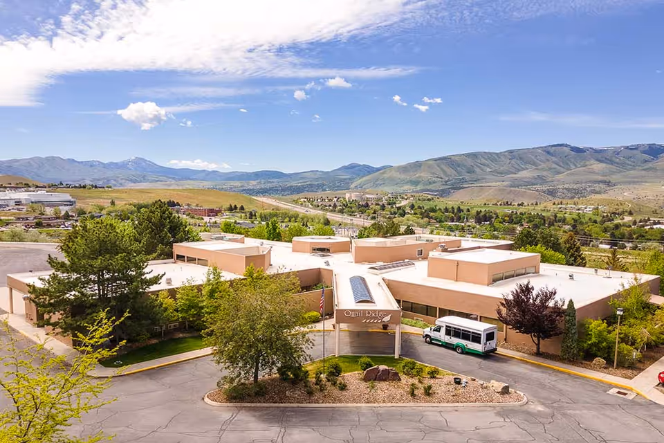 Aerial view of Quail Ridge Assisted Living & Memory Care facility surrounded by trees and landscaping, with mountains in the background under a partly cloudy blue sky. A white and green shuttle bus is parked near the entrance.