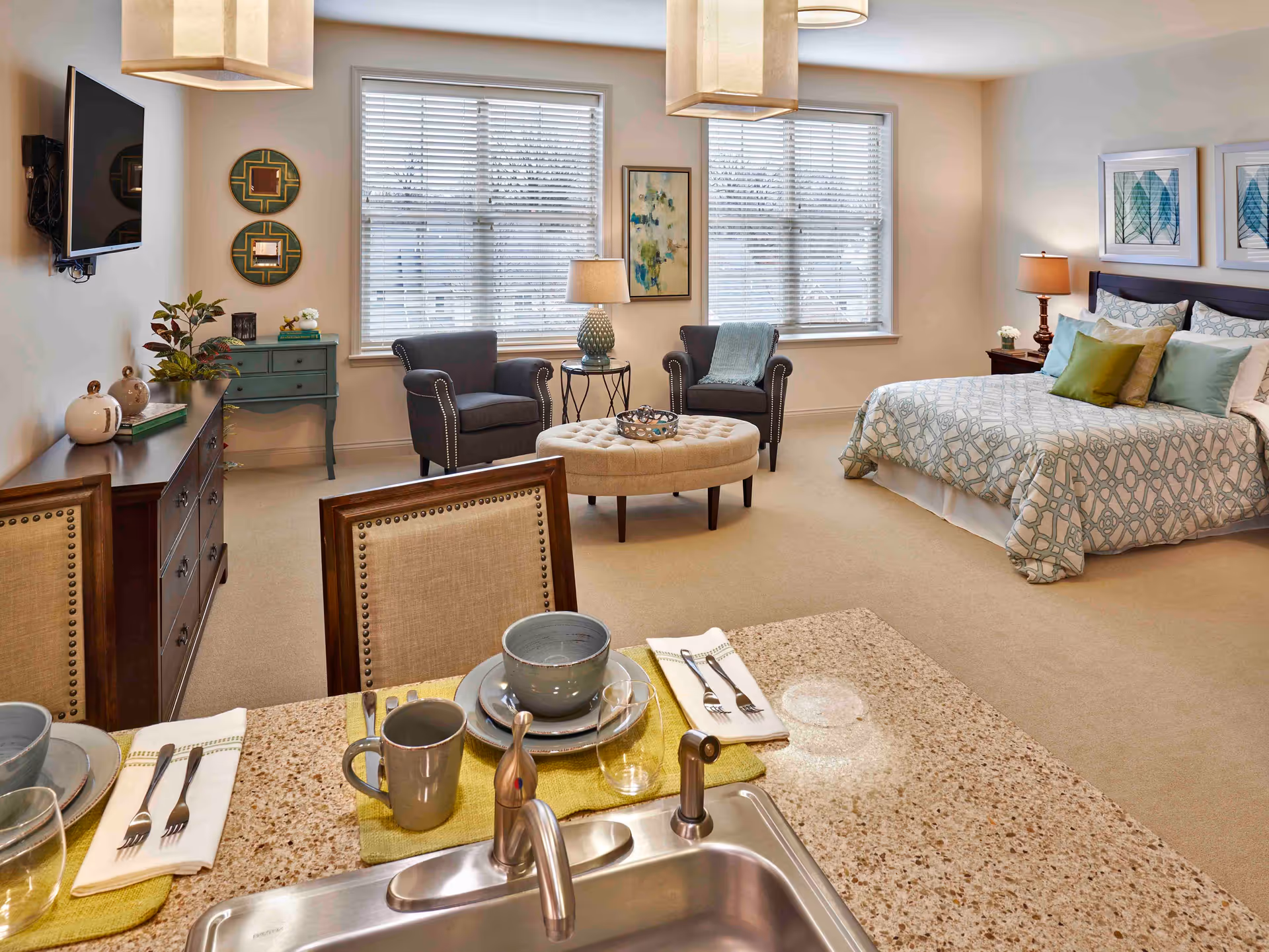 A spacious and well-lit senior living suite featuring a sleeping area with a bed adorned with patterned bedding and decorative pillows, a sitting area with two armchairs and a round tufted ottoman, a dining area with a table set with dishes and utensils, and a kitchen countertop with a sink and faucet. The room has large windows with blinds, framed artwork on the walls, and warm lighting from ceiling fixtures and table lamps.