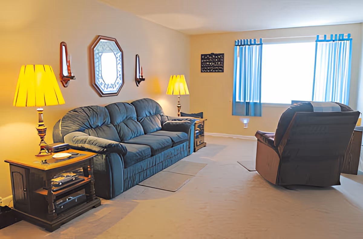 A cozy living room with a blue upholstered sofa and a matching armchair. There are two wooden side tables with lamps on either side of the sofa. A decorative mirror and two candle sconces hang on the wall above the sofa. A large window with blue curtains lets in natural light.