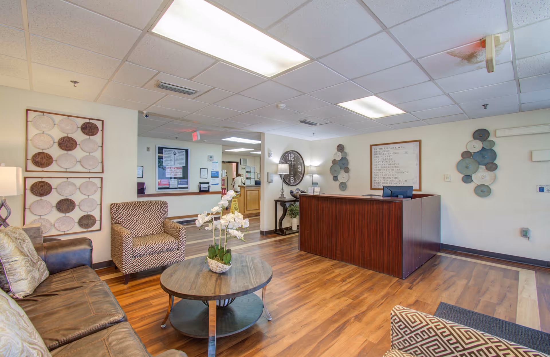 Reception lobby with a wooden front desk, seating area with chairs and a sofa, a round coffee table with flowers, and decorative wall art.