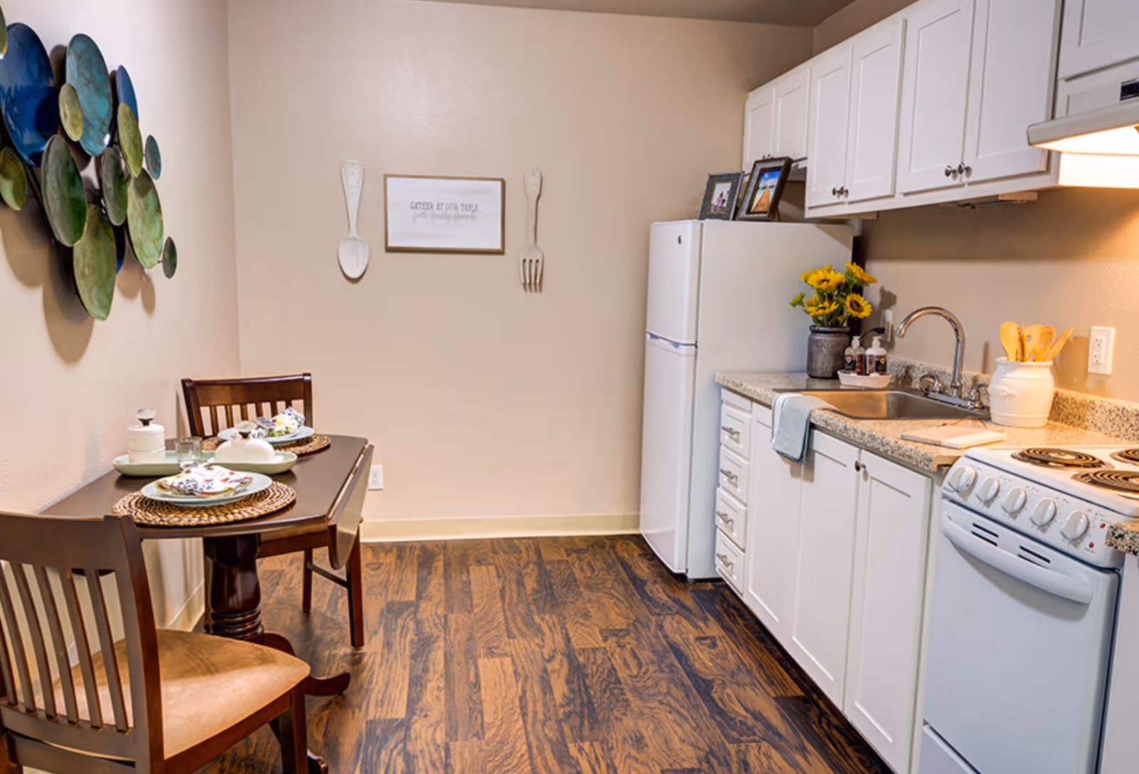 Small bright kitchen area with white cabinets, refrigerator, sink and stove, and a two-person dining table set against the wall.