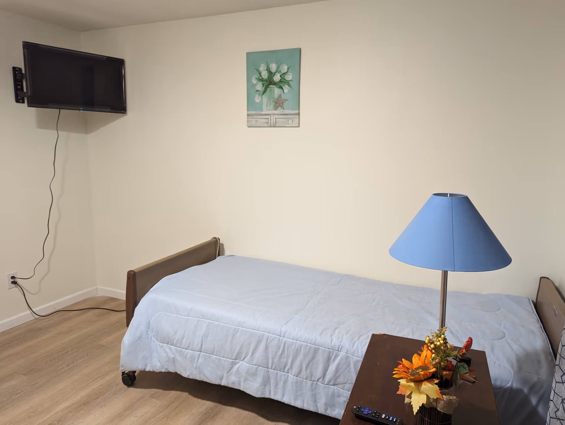 A simple bedroom with a single bed covered in a light blue comforter. A small wooden nightstand next to the bed holds a blue lampshade and a small decorative arrangement with orange flowers and autumn leaves. A flat-screen TV is mounted on the wall in the corner, with a visible power cord plugged into an outlet. A painting of white flowers in a vase hangs on the wall above the bed. The floor is light wood, and the walls are painted beige.
