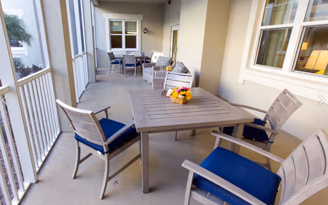 Covered outdoor patio with a square table topped by a small flower basket, surrounded by four chairs with blue cushions and additional seating along the wall.