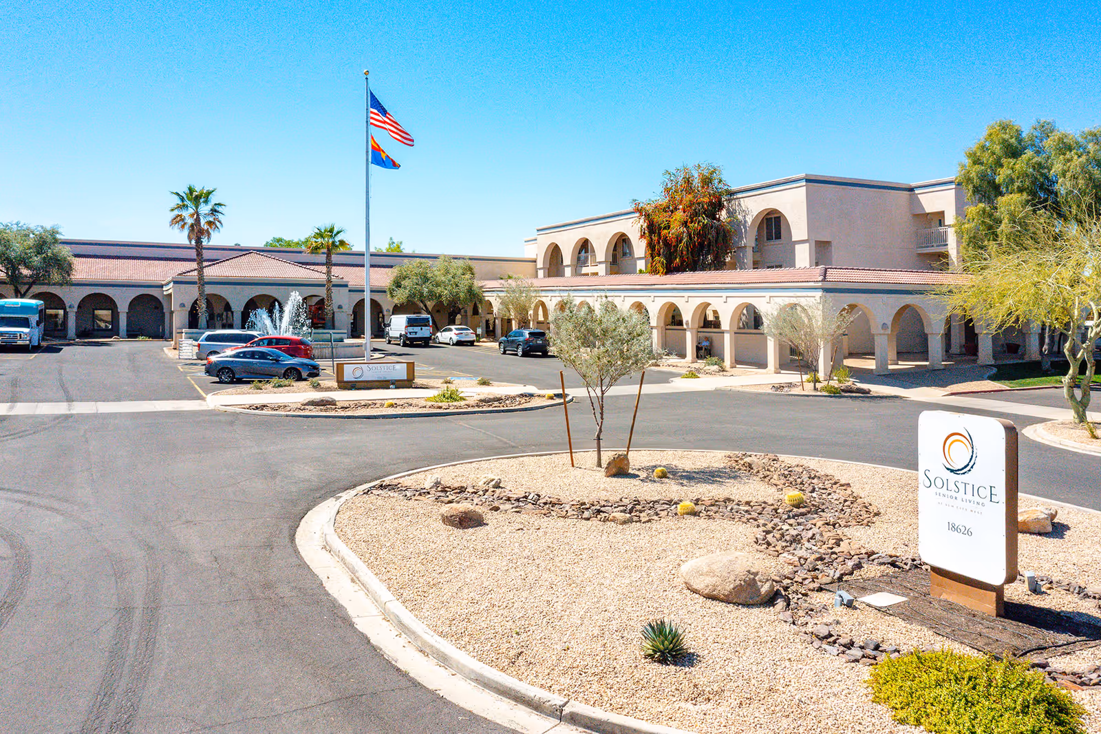 Exterior view of Solstice Senior Living at Sun City West showing a large building with arches, a parking lot with several cars, a fountain, two flagpoles with the American and Arizona state flags, and desert landscaping with rocks and small plants under a clear blue sky.