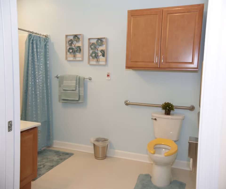A clean and accessible bathroom with light blue walls, a white toilet with a yellow seat, a wooden cabinet mounted on the wall above the toilet, a metal grab bar beside the toilet, a small green plant on the toilet tank, a blue shower curtain, matching blue bath mats, a towel rack with folded blue towels, two framed floral wall decorations, and a small trash can on the floor.