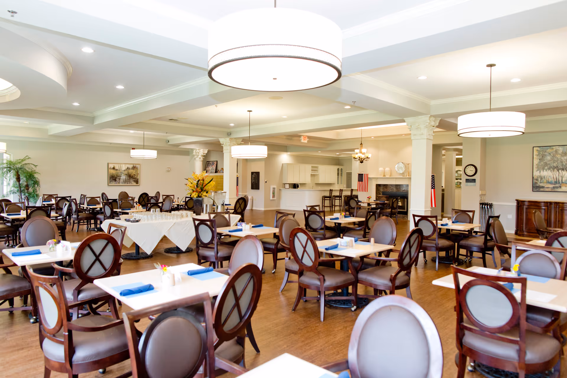 A spacious dining room with multiple tables and chairs arranged neatly. Each table is set with blue napkins and small flower vases. The room features large round ceiling lights, a fireplace, and decorative columns. There are paintings on the walls and an American flag near the fireplace.