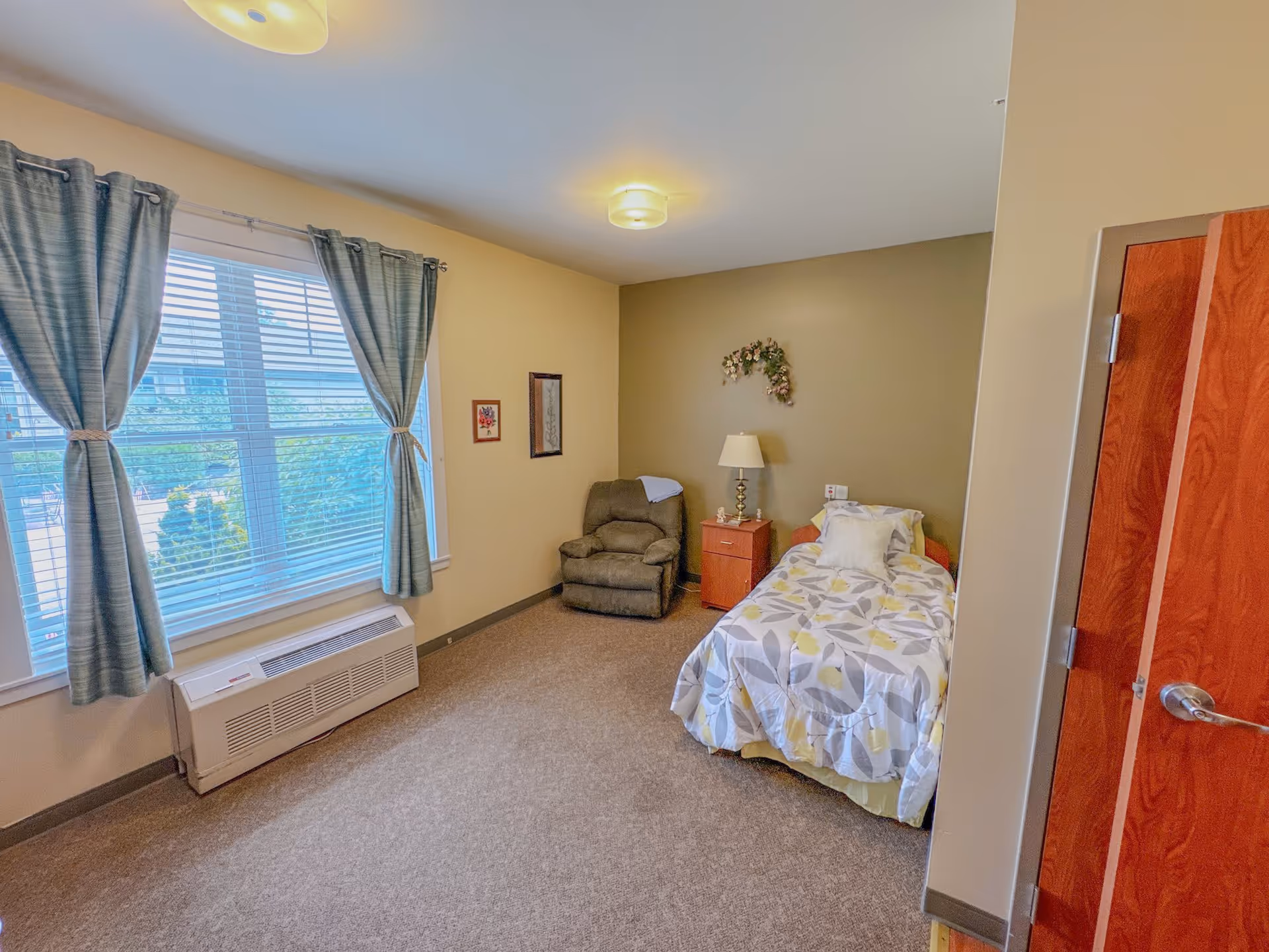 A cozy bedroom in a senior living facility with a single bed covered in a floral patterned comforter, a wooden nightstand with a lamp, a comfortable armchair, and a large window with blue curtains letting in natural light.